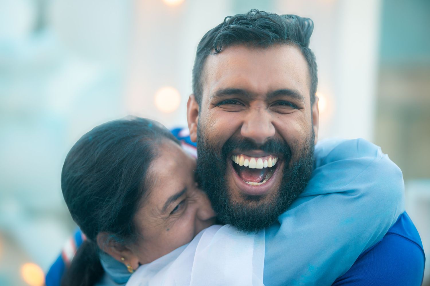 Happy bearded man laughing while receiving warm hug from older woman at outdoor family gathering.