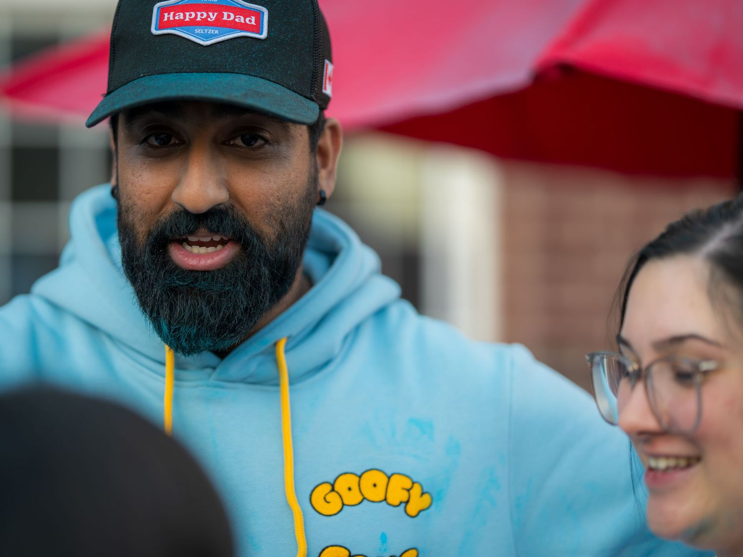 Man wearing Happy Dad cap and Goofy blue hoodie speaking at an outdoor gender reveal event.