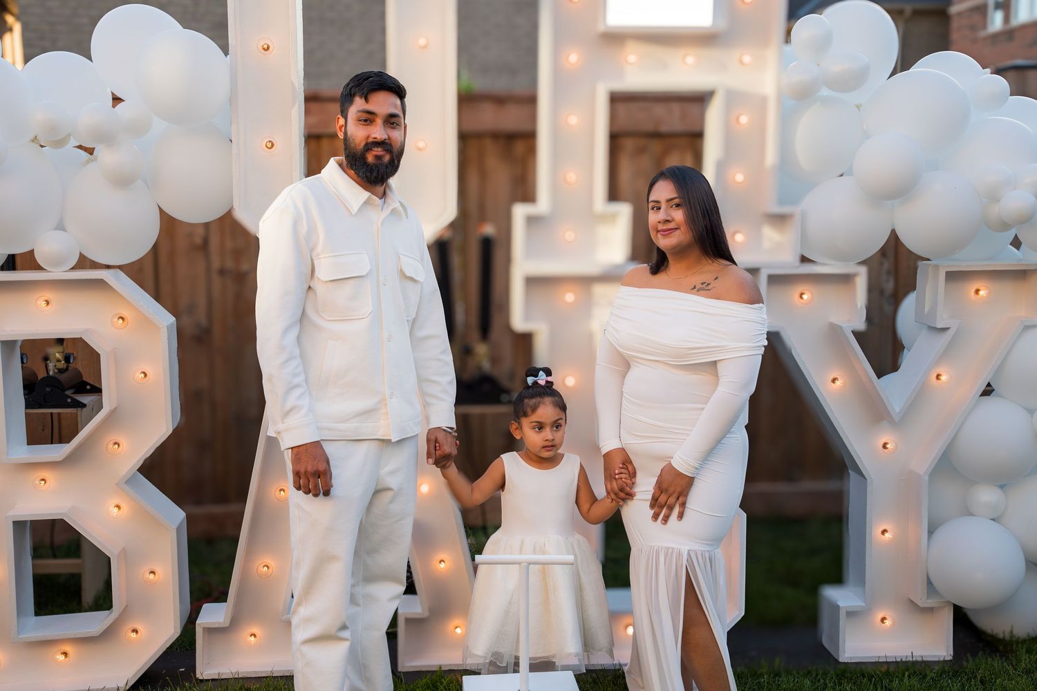 A couple in white holds their young daughter's hands beside a lit BABY marquee sign.
