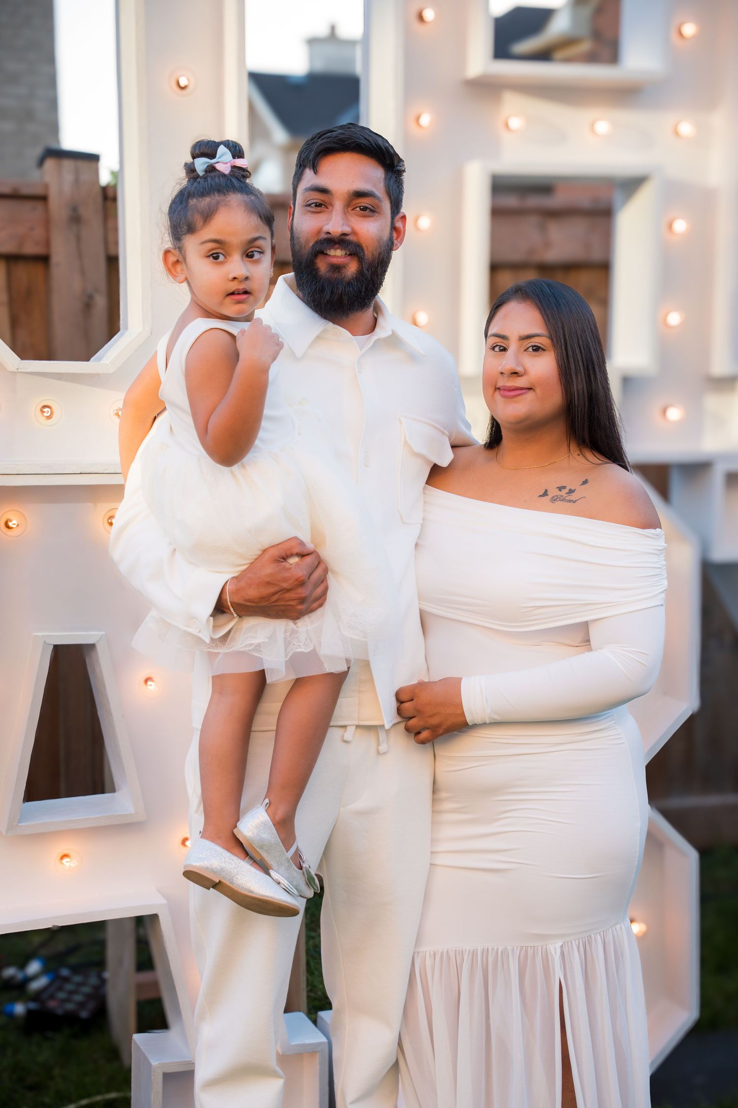 Bearded man holds young daughter while standing beside pregnant woman in white dress at baby shower.