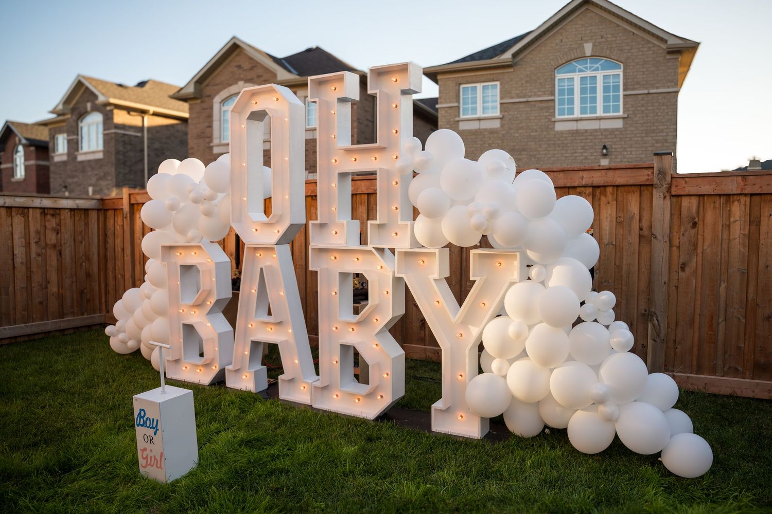 Illuminated OH BABY marquee letters surrounded by white balloon garland arch at an outdoor gender reveal party.