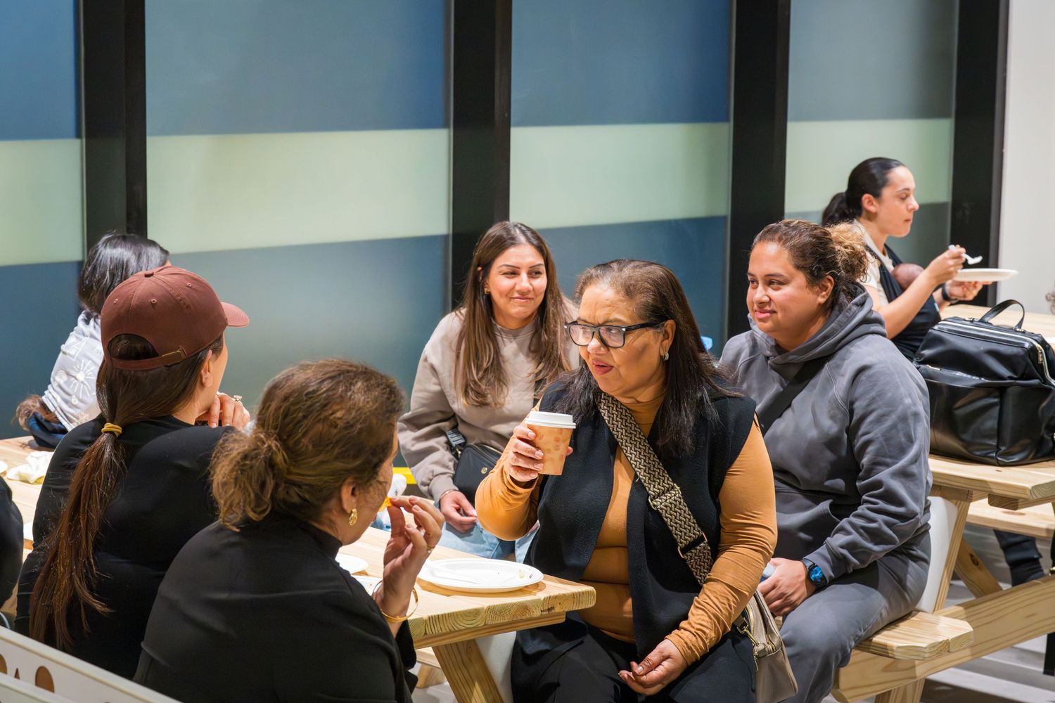 Group of women smiling and chatting together at a picnic table in a bright indoor community space.
