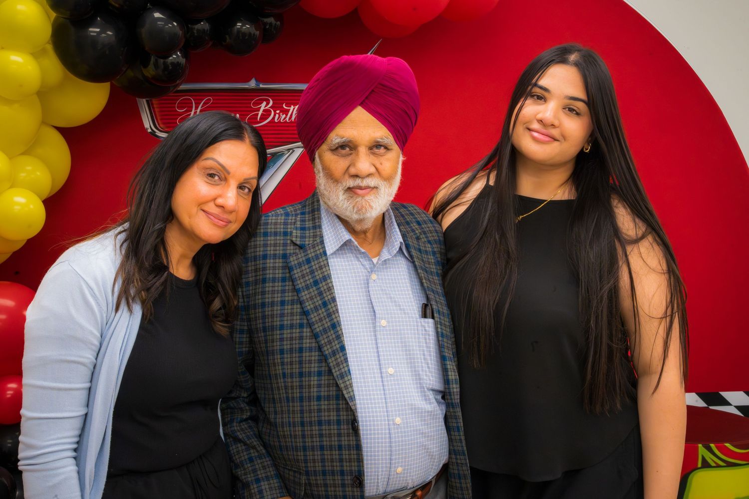 Three people smiling together at a festive celebration with red and yellow balloon decorations in the background.