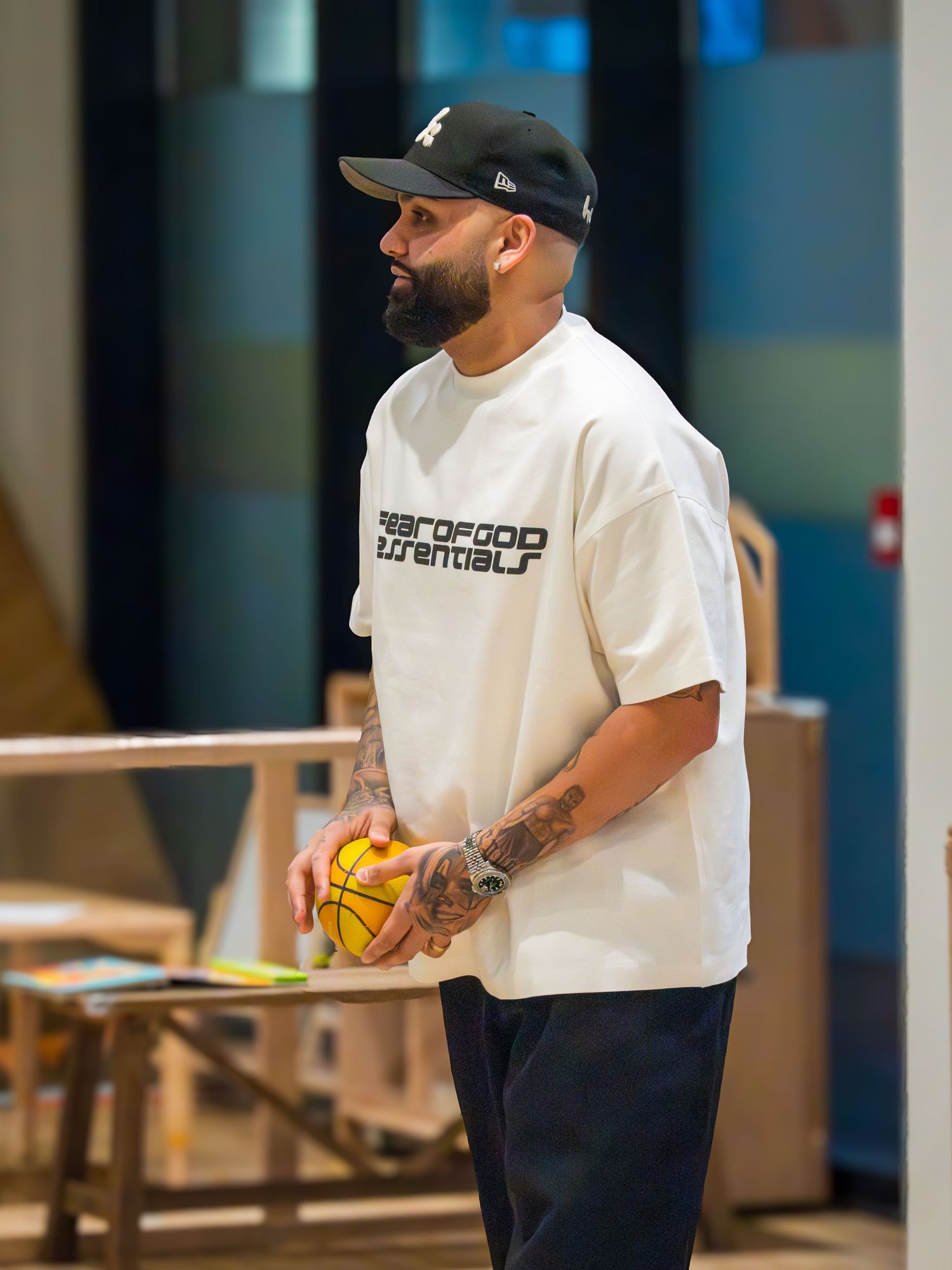 A bearded tattooed man in a white Fear of God tee holds a yellow ball at an indoor play space.