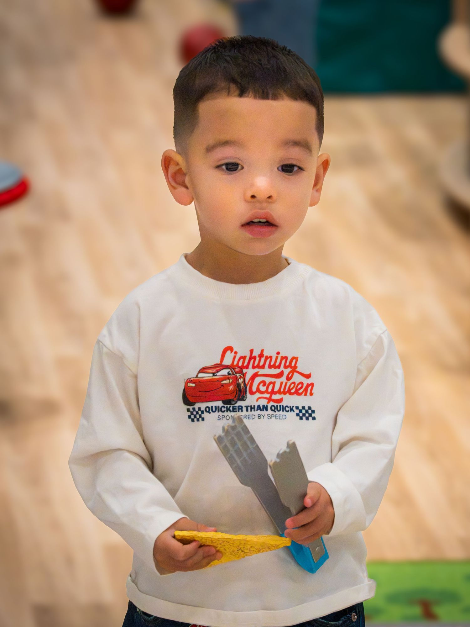 Young toddler boy wearing a Lightning McQueen white sweatshirt holding toy blocks in a playroom.