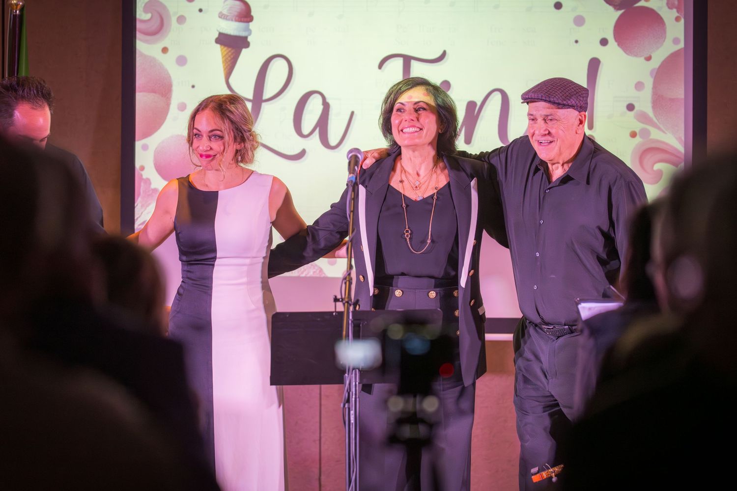 Three performers smile and embrace on stage in front of a La Fine projected backdrop at show end.