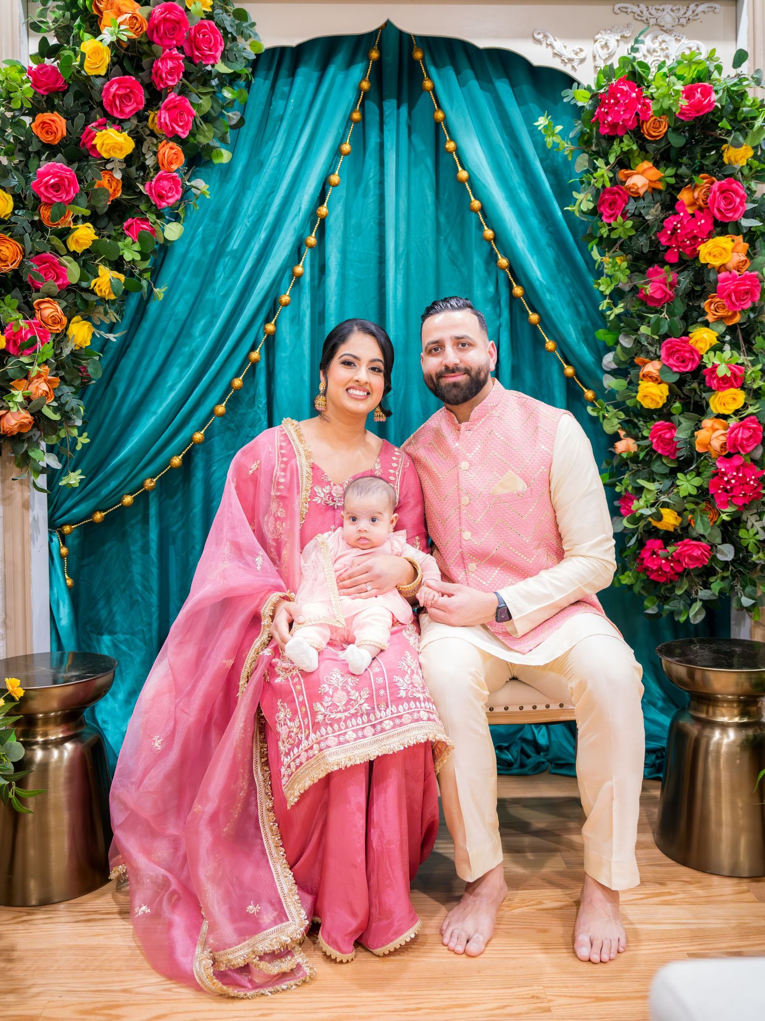 Indian family portrait with couple in pink and cream traditional attire holding baby before teal floral backdrop.