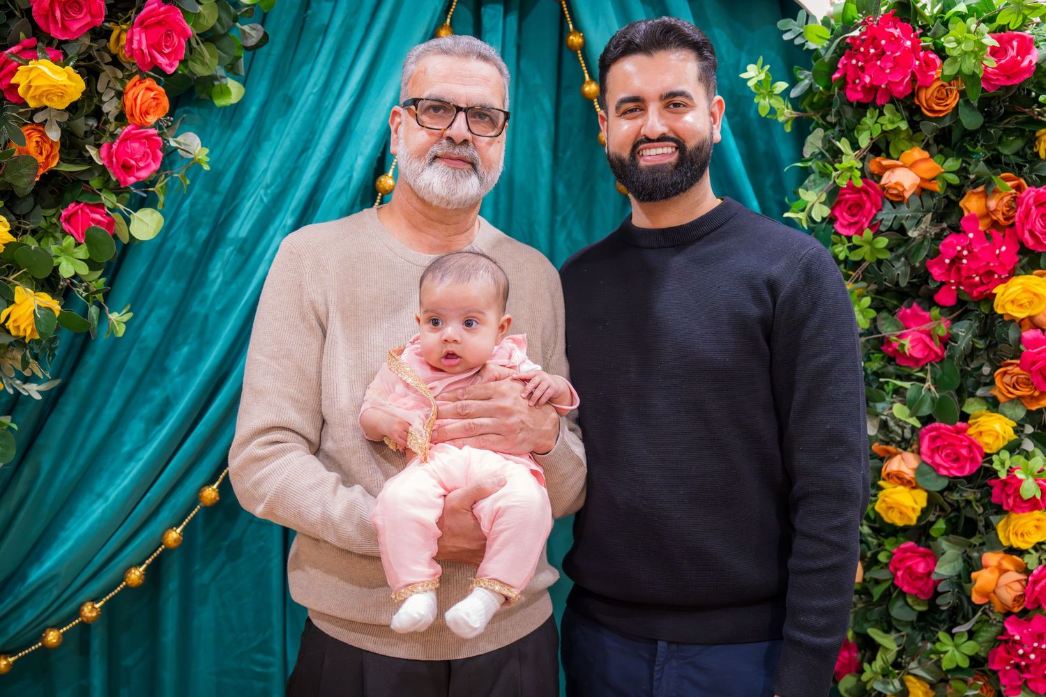 Two men pose with a newborn baby dressed in pink in front of a vibrant floral and teal curtain backdrop.