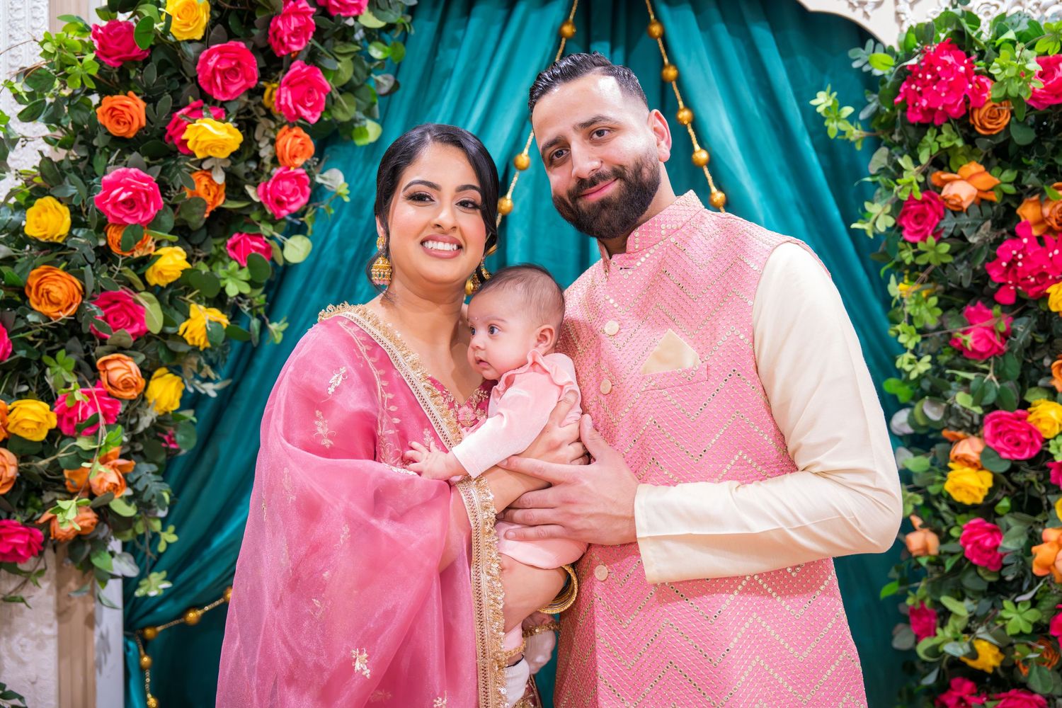Happy South Asian family with newborn baby posing before vibrant flower arch backdrop.