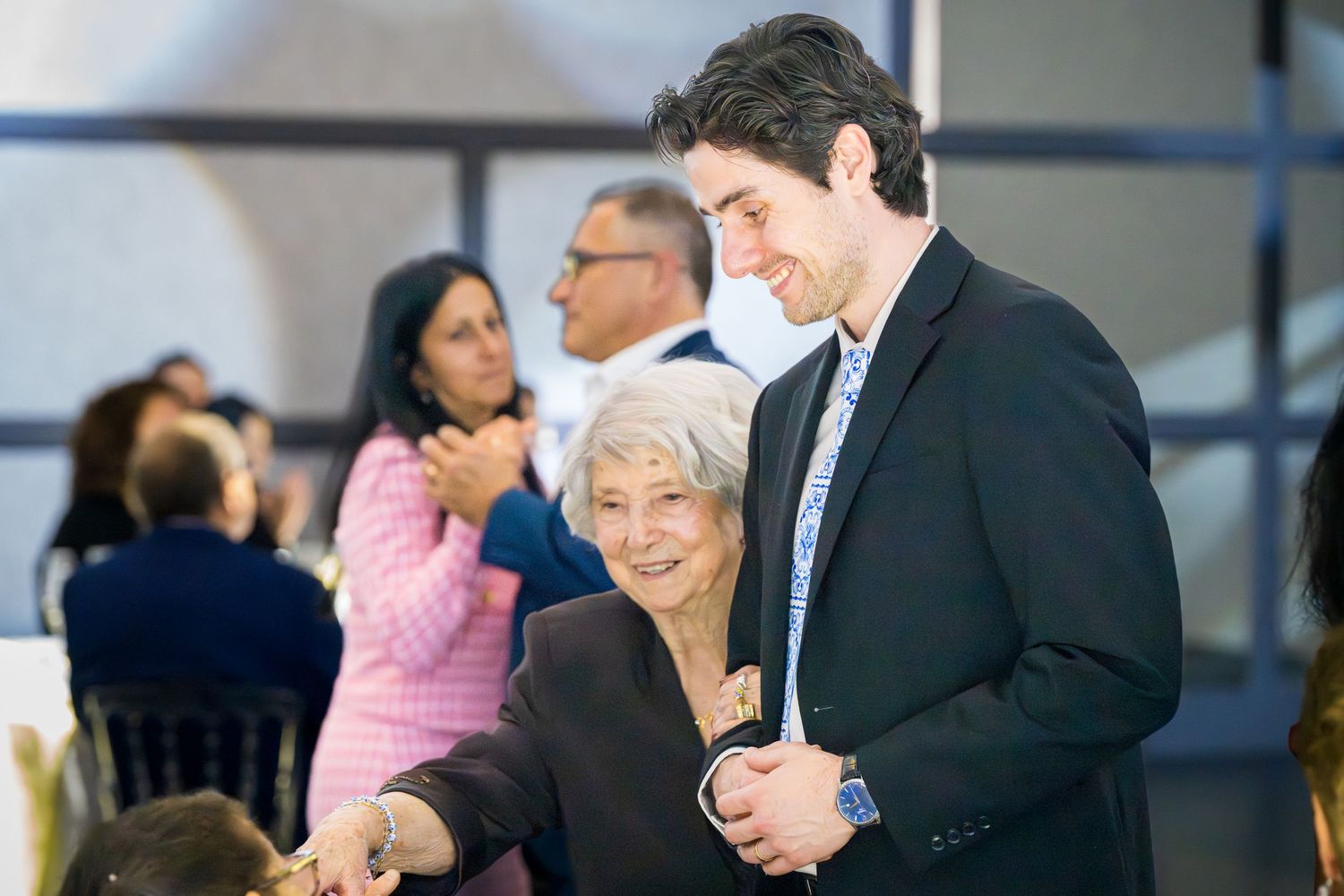 Young man smiling and embracing an elderly woman at a formal celebration event indoors.