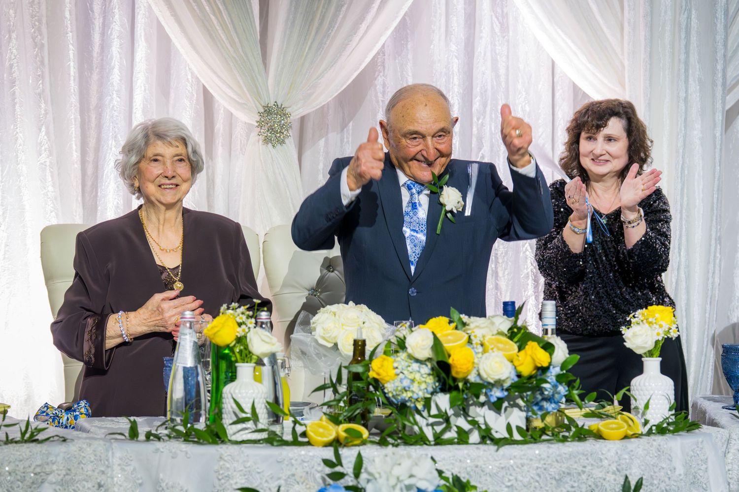 Three people smiling and celebrating at an anniversary party head table with yellow and white floral arrangements.