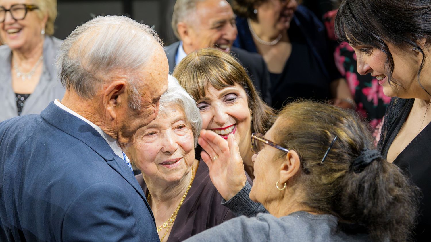 Elderly man warmly greeting smiling women at a social gathering event.