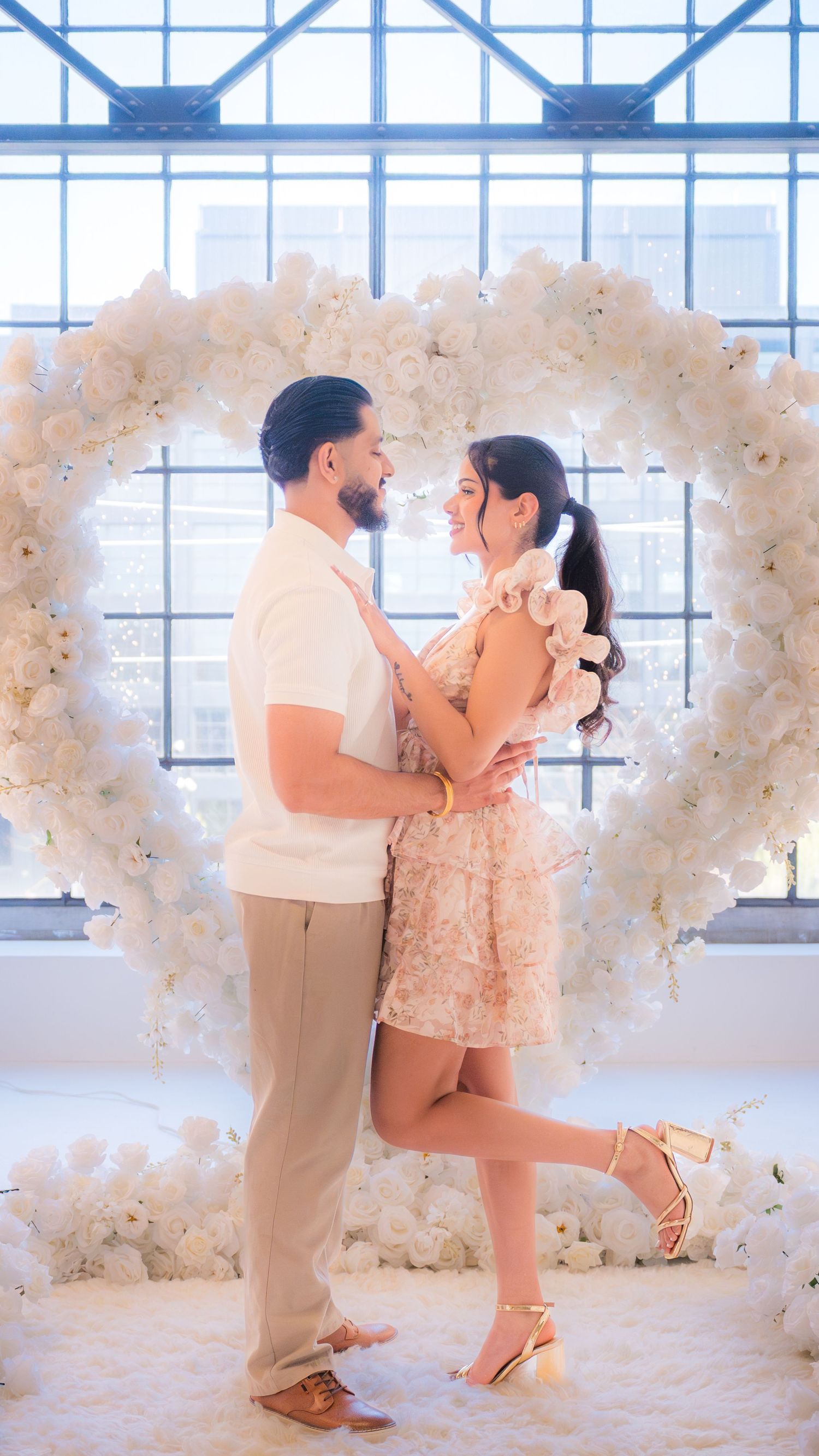 Happy couple embracing in front of white floral heart arch during romantic marriage proposal.