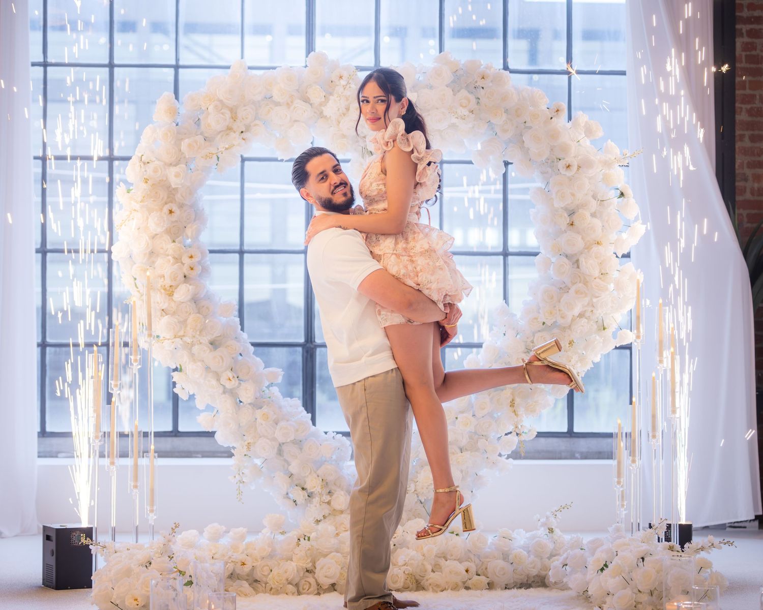 Happy couple posing in front of a white floral heart arch with sparklers at an engagement celebration.