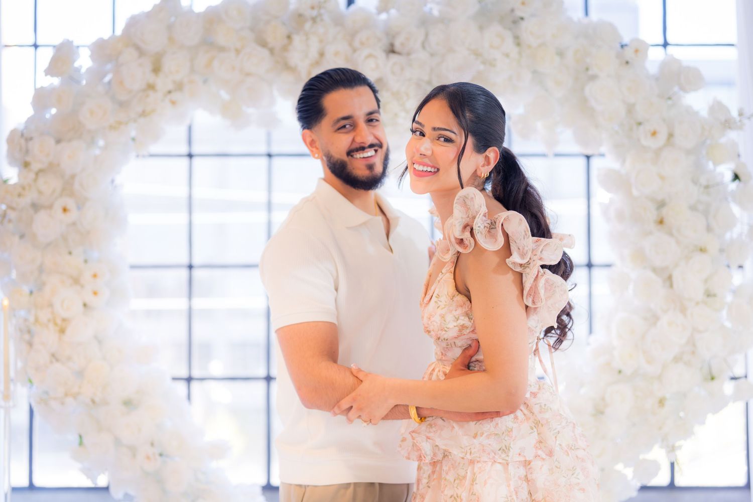 Engaged couple posing together inside a stunning circular white floral arch for a proposal photo.