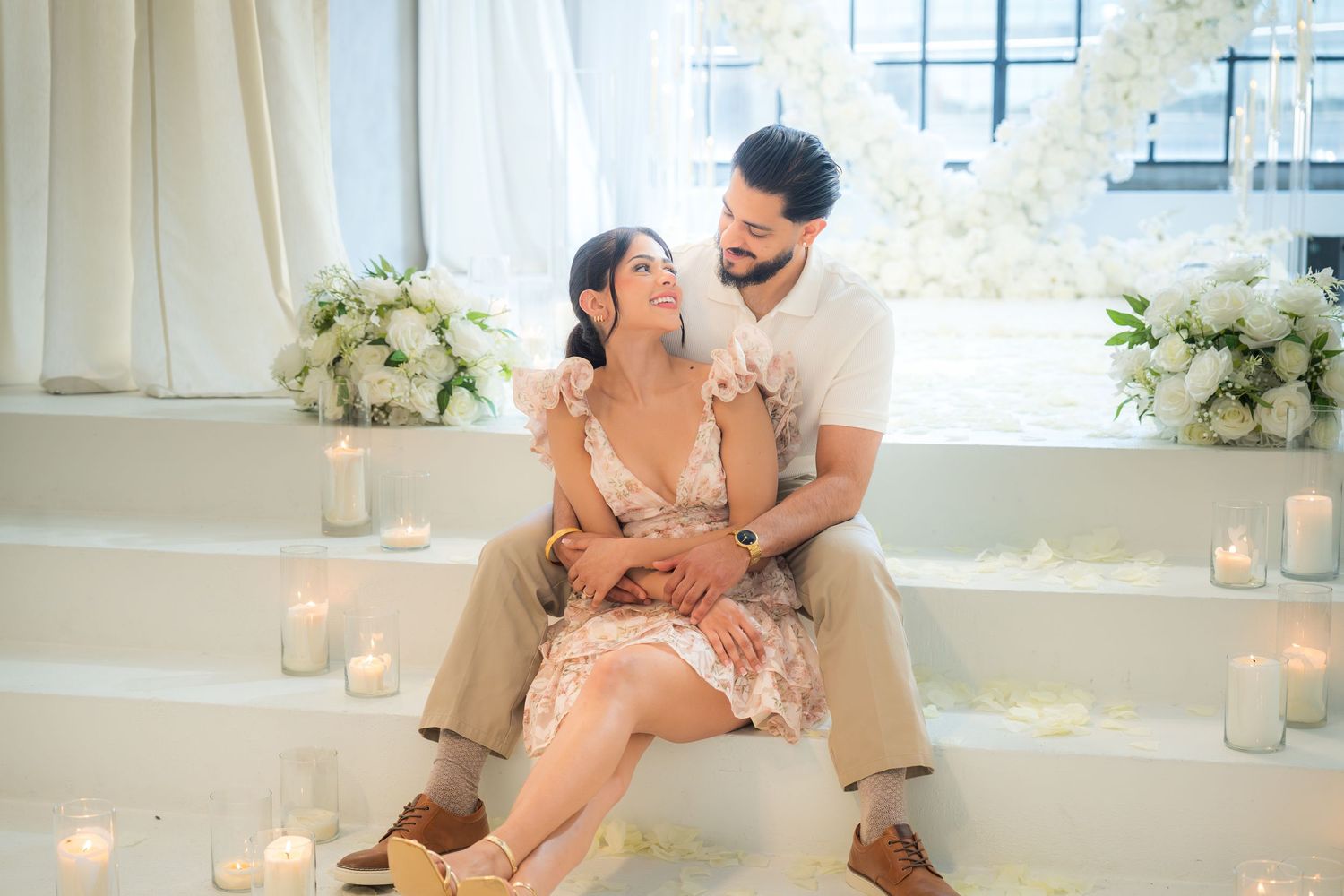 Engaged couple sitting together on steps surrounded by candles and white rose petal decorations.