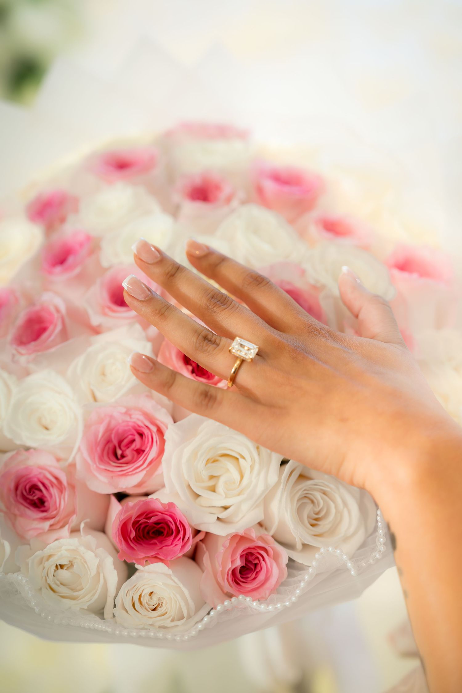 Close-up of hand wearing gold diamond engagement ring resting on pink and white rose bouquet.
