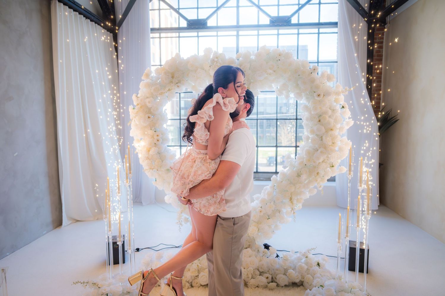 Newly engaged couple embracing and kissing in front of a glowing white floral heart arch with fairy lights.
