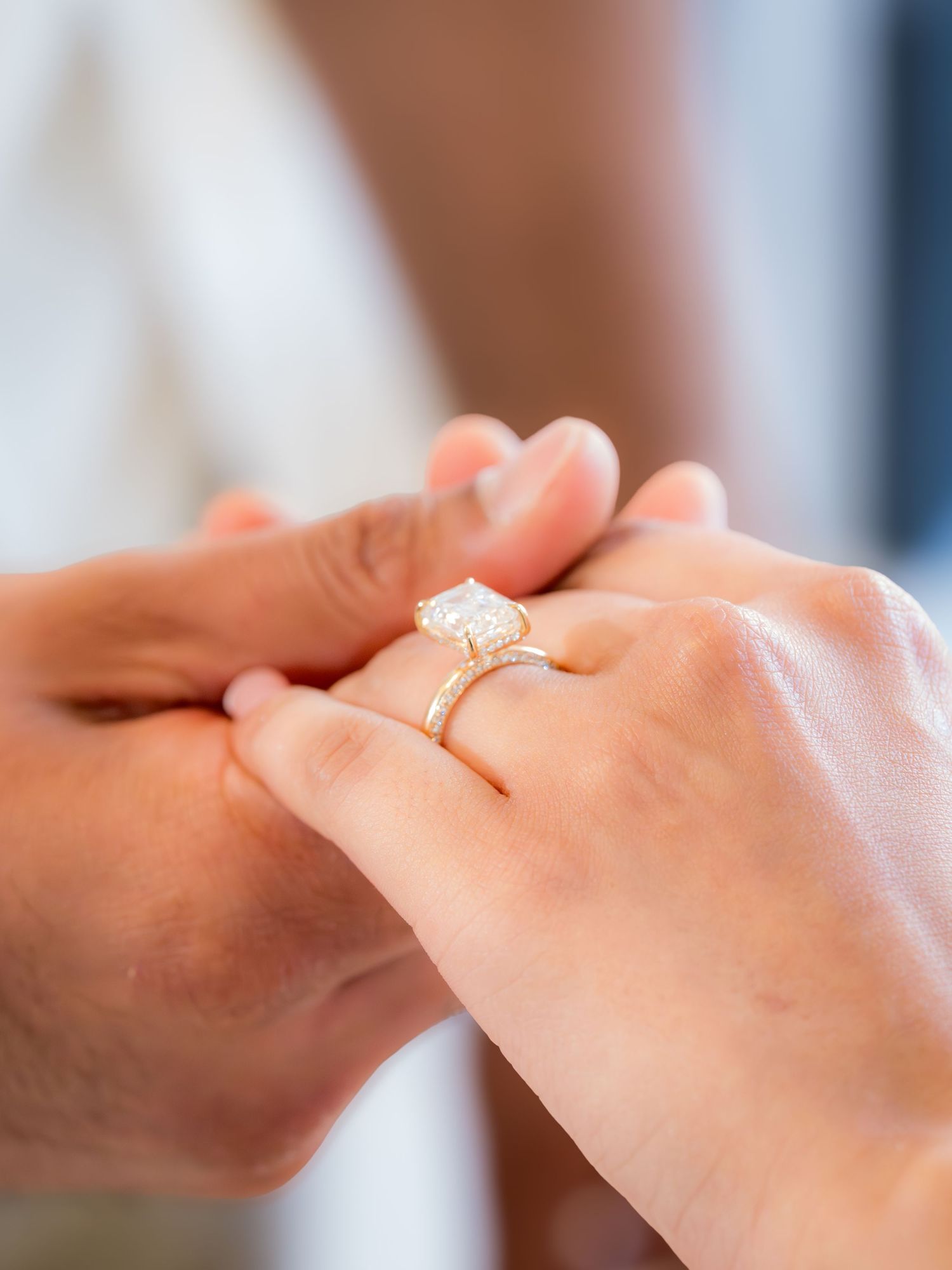Close-up detail of emerald cut diamond engagement ring on gold band held by couple.