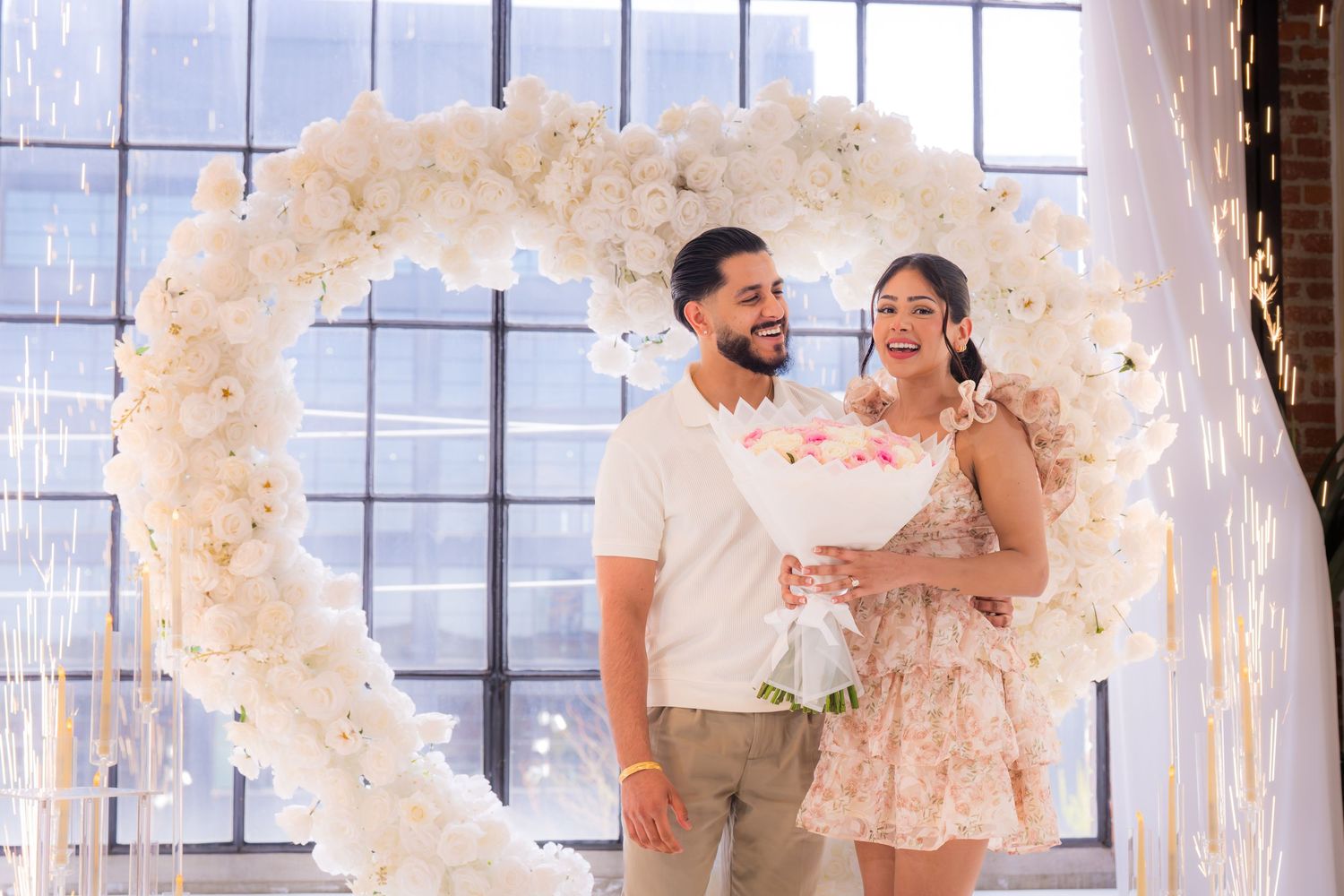 Newly engaged couple smiles together holding a bouquet in front of a white floral heart arch.