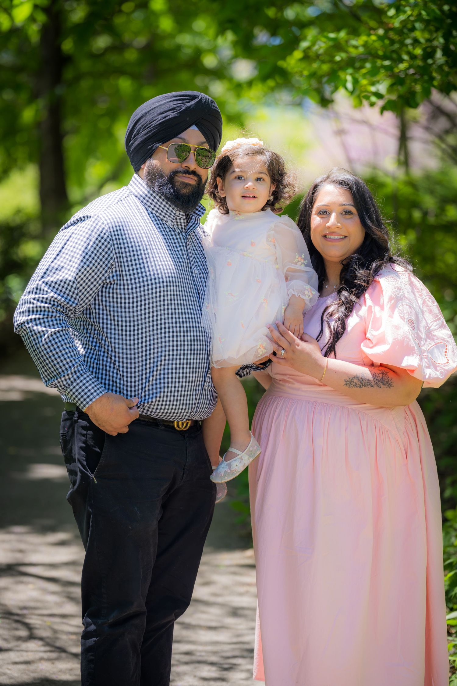 A Sikh family poses outdoors, with a man in a turban holding a toddler beside a pregnant woman in pink.