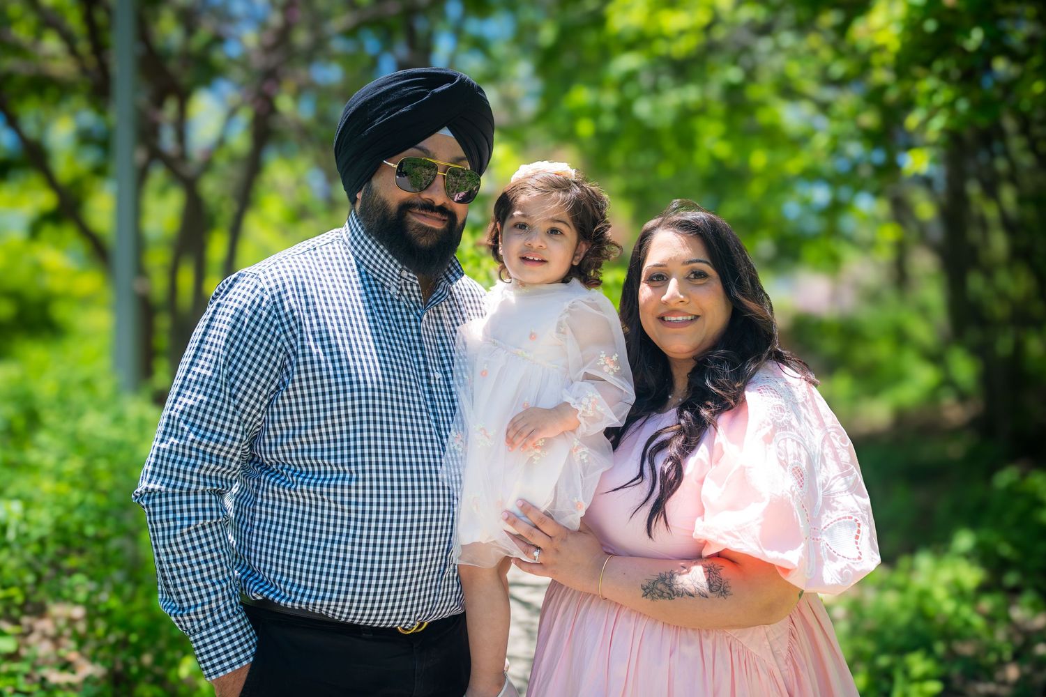 A Sikh family with a man in turban, woman in pink dress, and toddler in white posing outdoors.