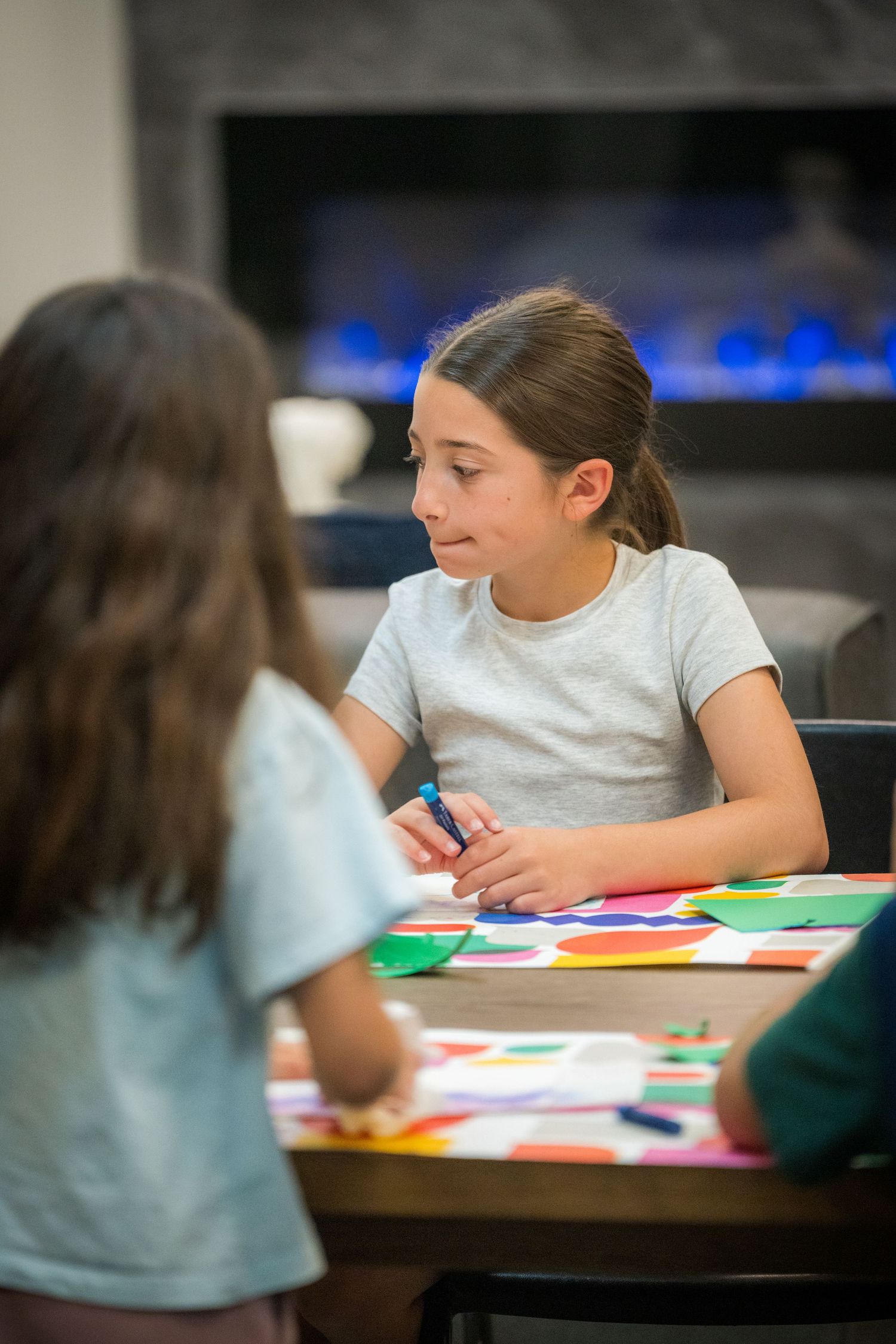 Girl in gray t-shirt holds marker while doing colorful art project at table with fireplace in background.