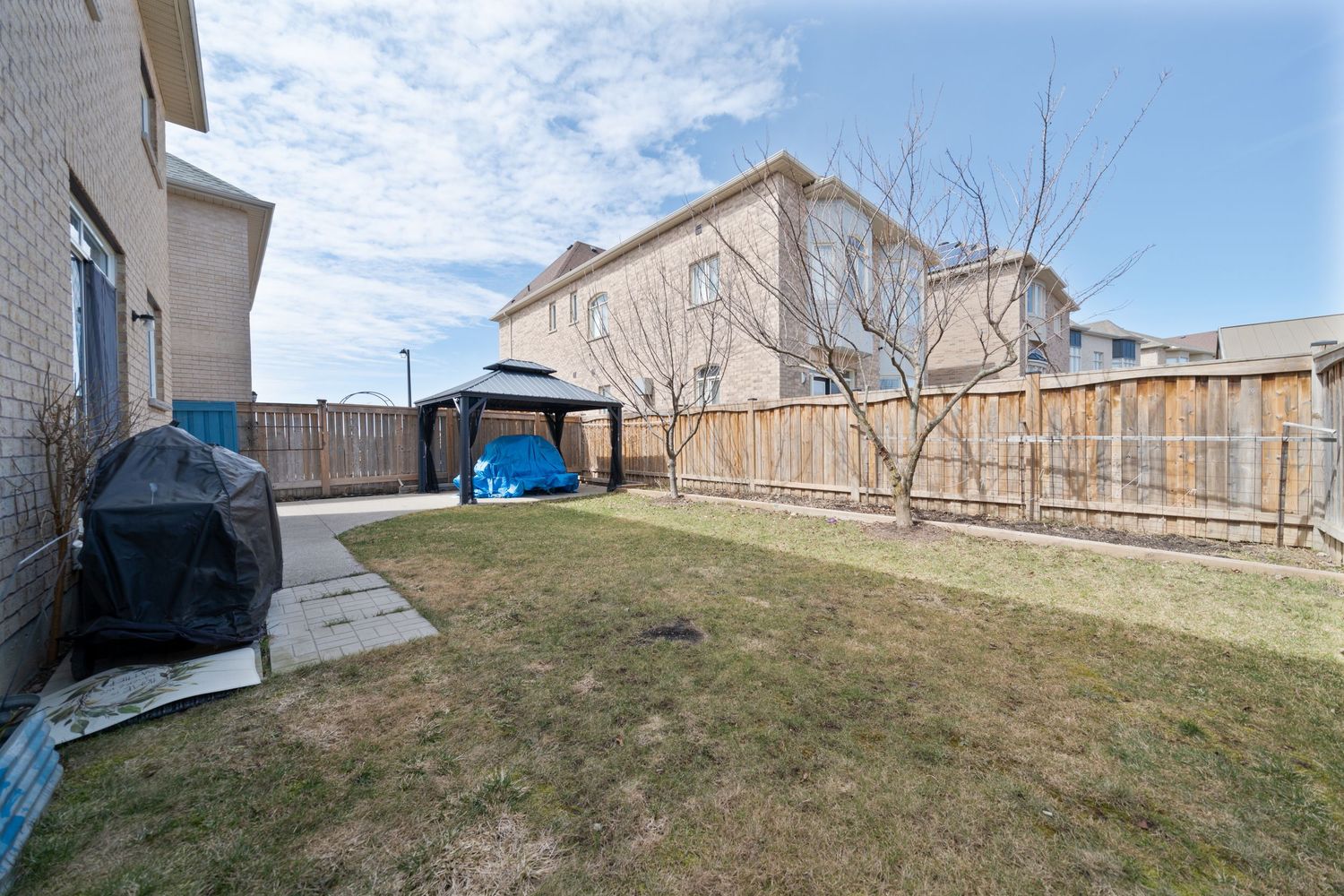 Fenced backyard with brown grass, wooden privacy fence, and a covered barbecue grill against a residential house.