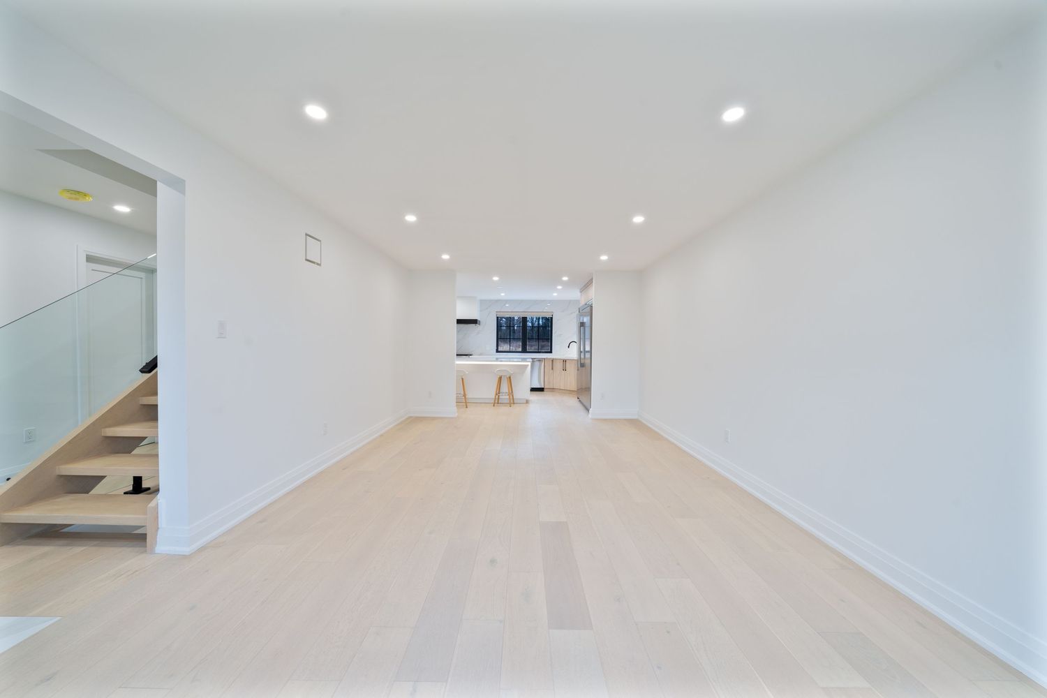 Modern white hallway with recessed lighting and hardwood flooring leads to a bright open space.