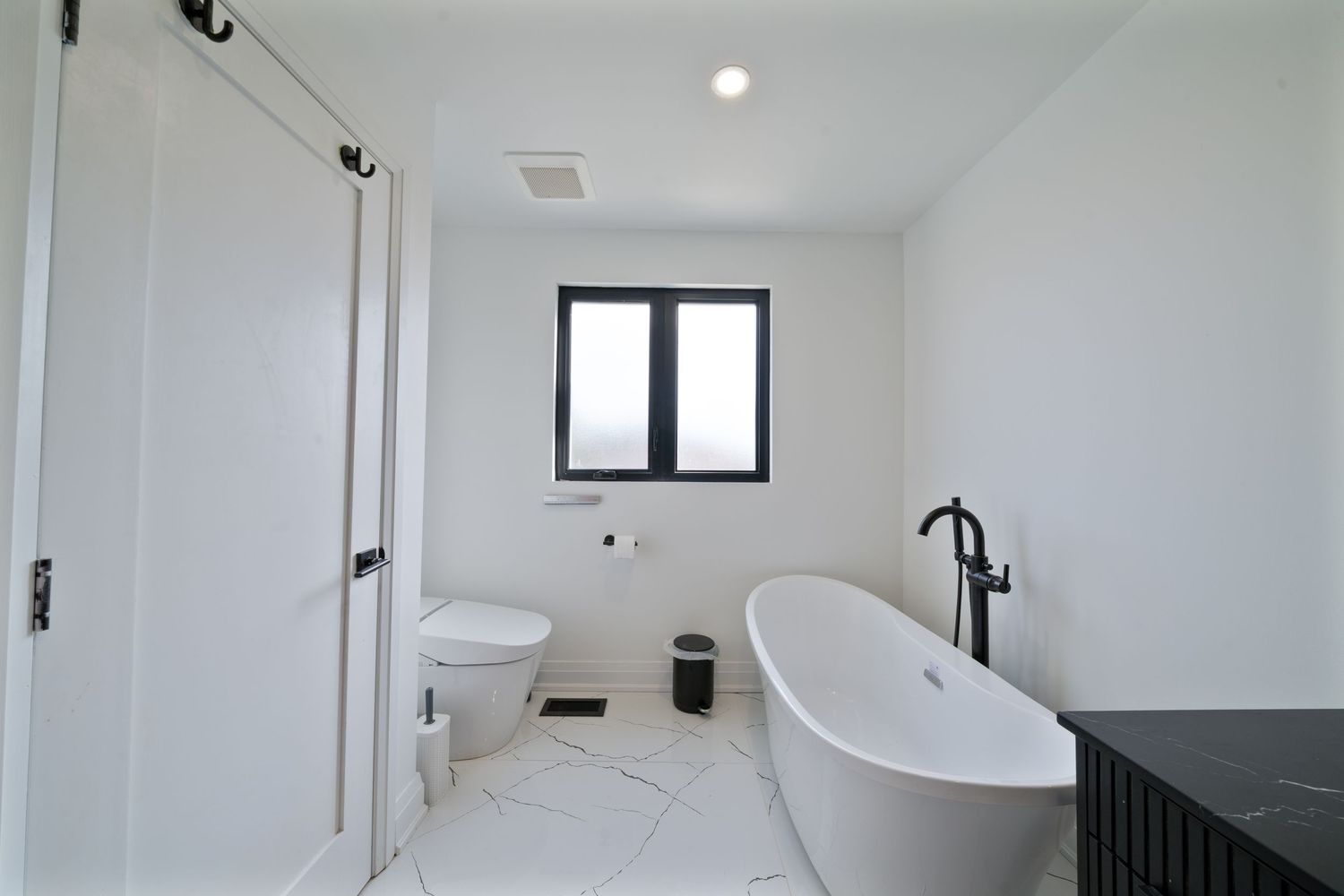 Modern bathroom interior with white walls, black fixtures, and a freestanding bathtub next to a window.
