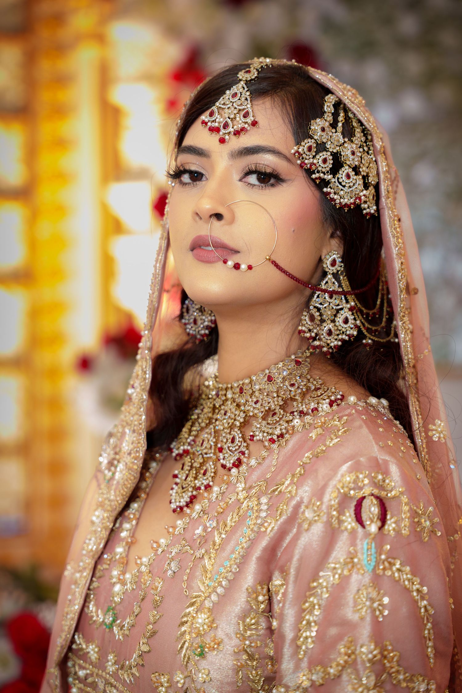 Ornate bridal jewelry and makeup details in soft pink and gold tones with floral backdrop.