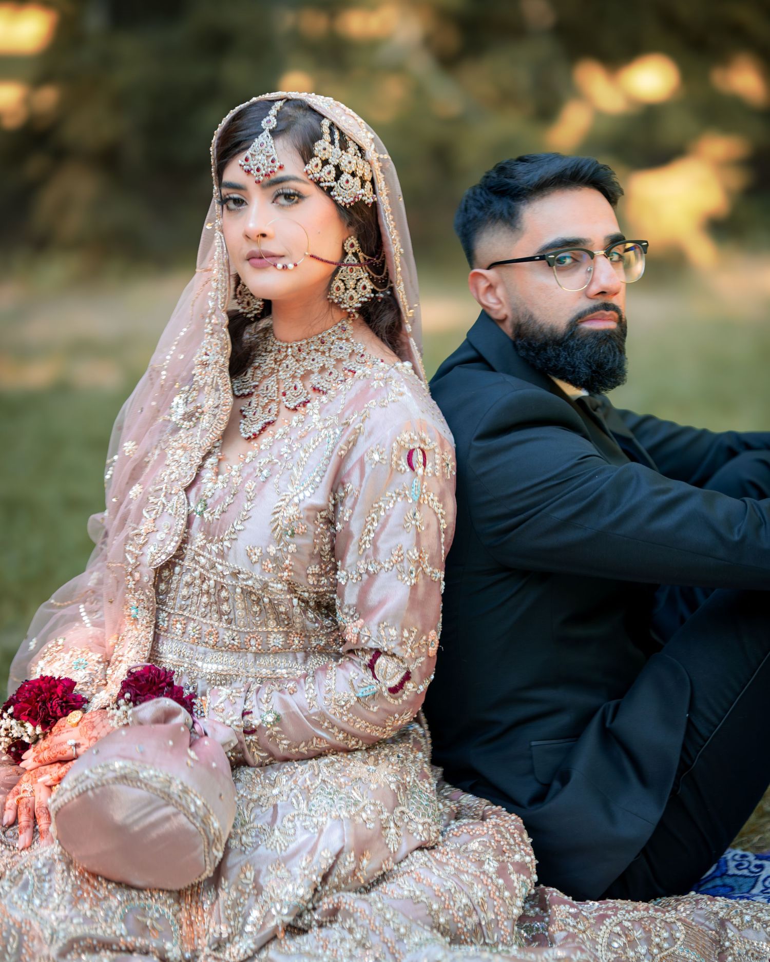 Couple in traditional South Asian wedding attire poses on grass during golden hour photography session.