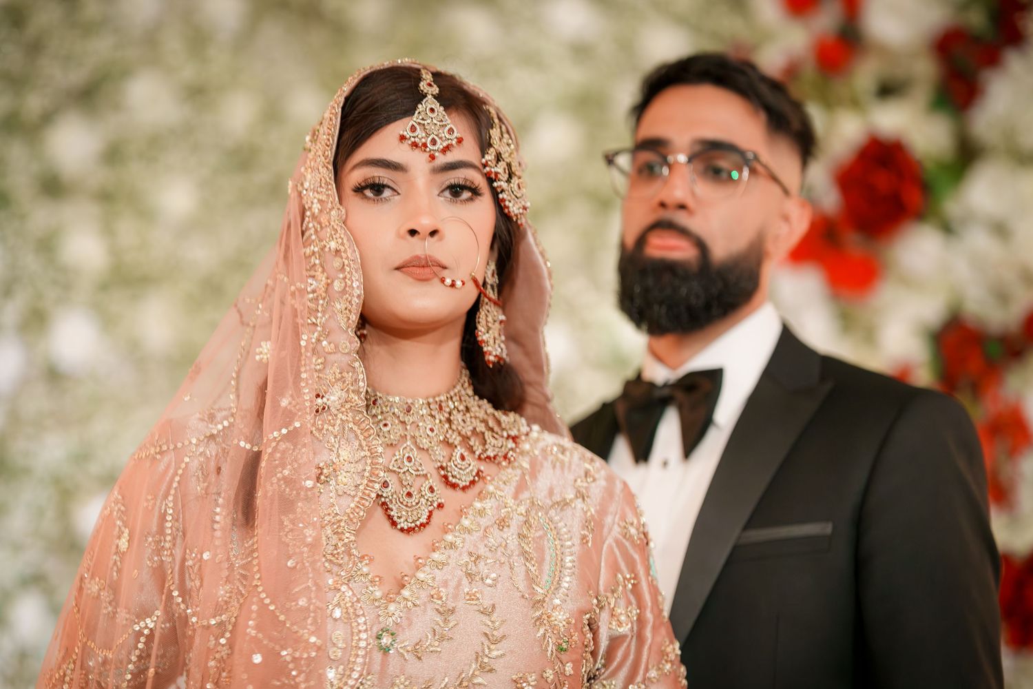 Couple in elegant South Asian wedding attire pose against floral backdrop during ceremony.