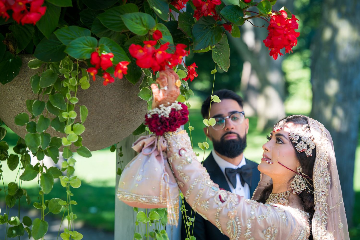 A couple poses in a garden setting with vibrant red flowers and lush greenery for wedding photos.