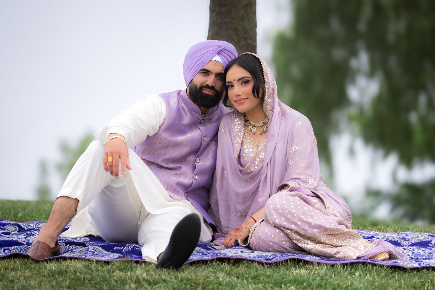 A group in coordinated purple and white traditional clothing poses together on a decorative blanket outdoors.