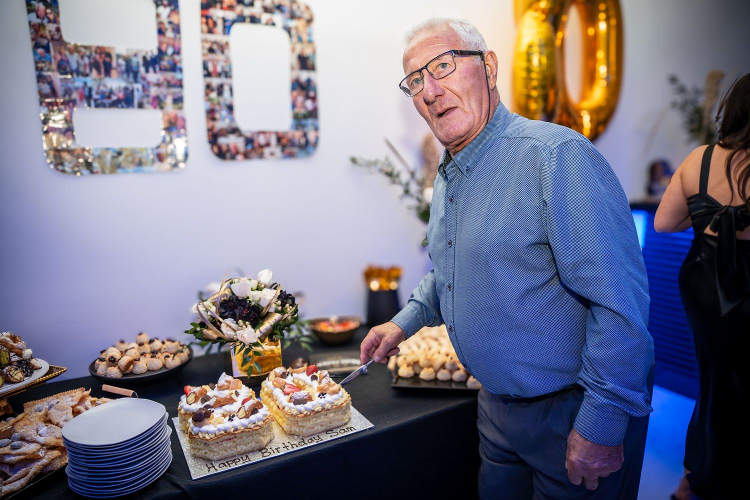 An older gentleman in glasses stands near a dessert table with cakes and treats.
