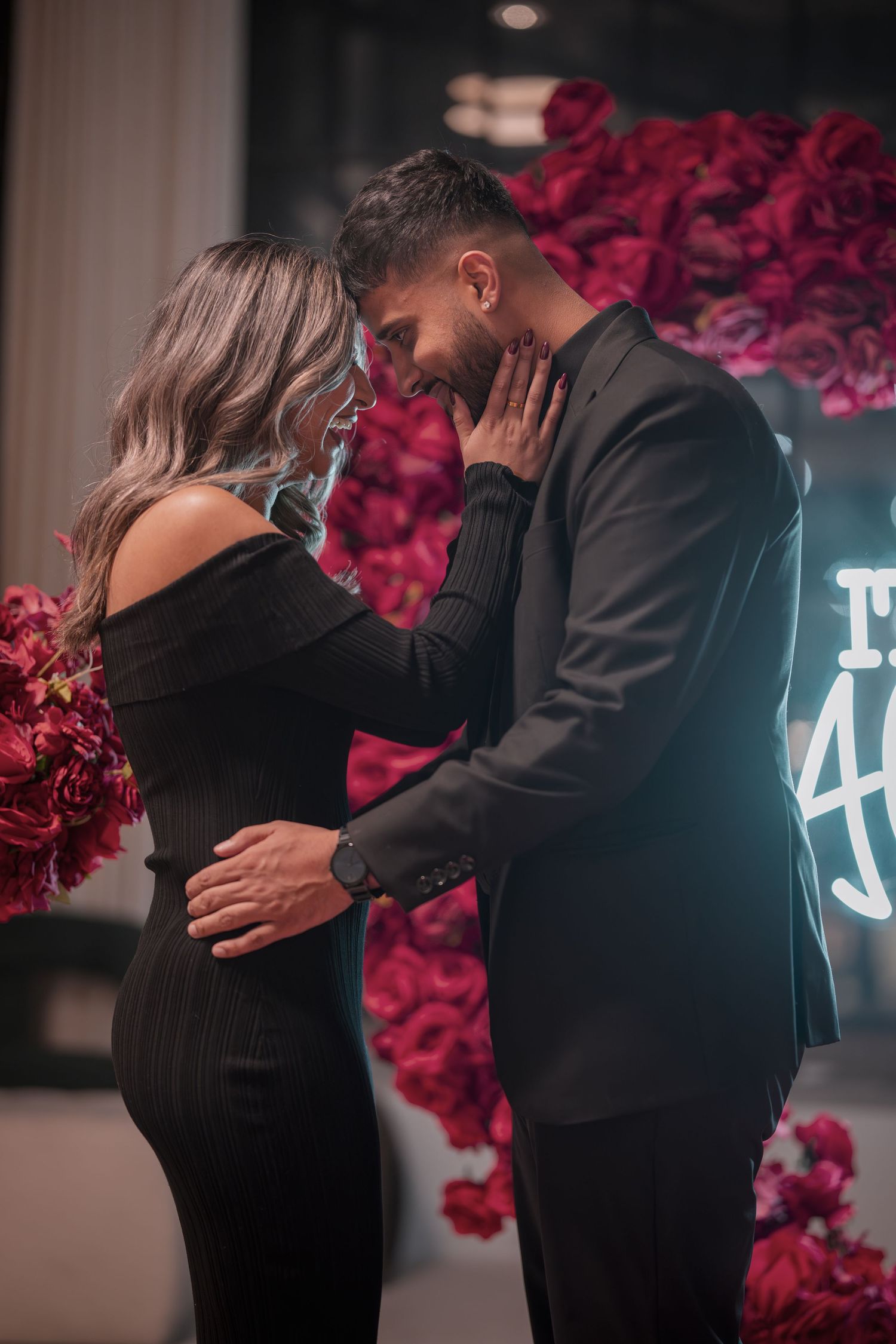 Intimate embrace captured in front of romantic red rose wall and neon lighting.