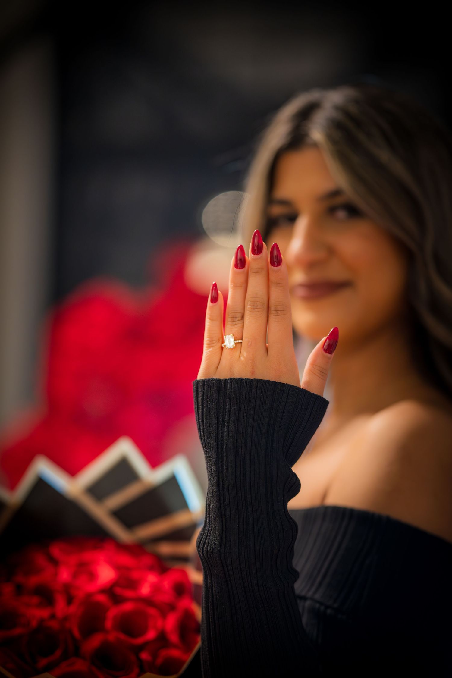 Woman showing off engagement ring with red manicure against backdrop of roses.