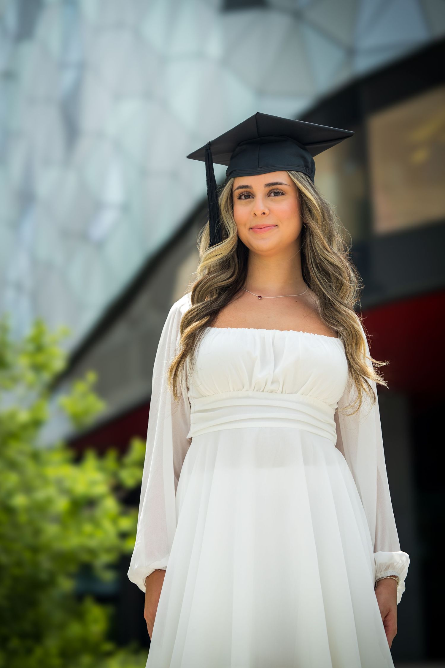 Graduate in white dress and cap poses outdoors for graduation photos against modern building.