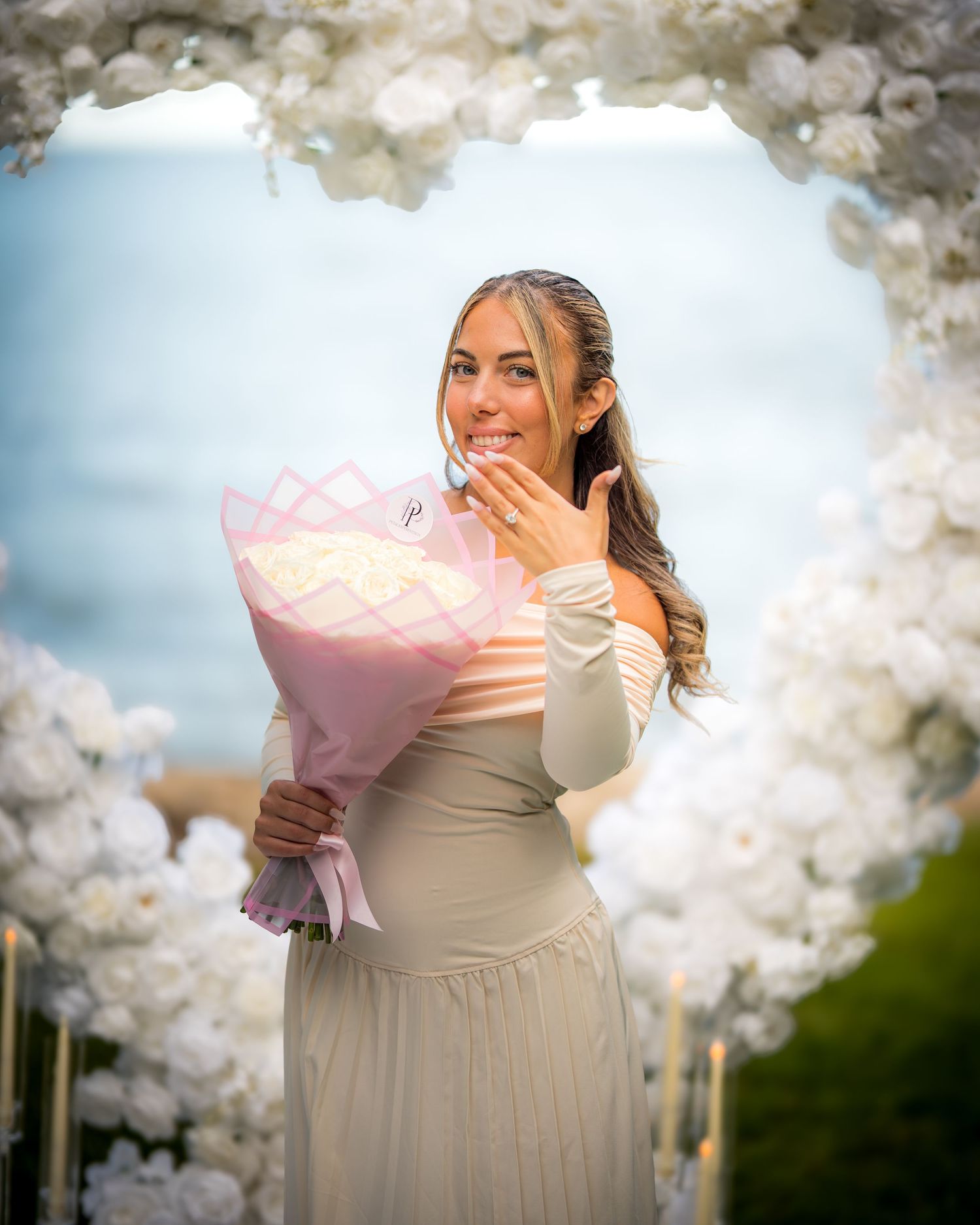 Person in cream dress holds bouquet while standing under white floral decorations outdoors.