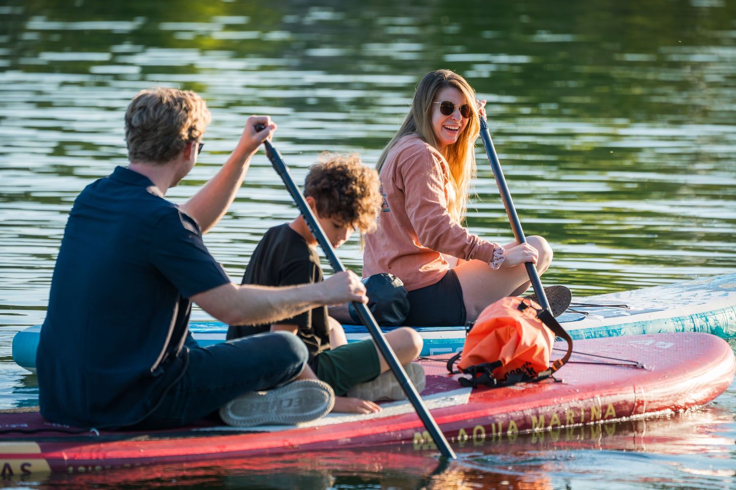 Three people paddleboarding on a calm lake, smiling and paddling together on a red inflatable SUP board.