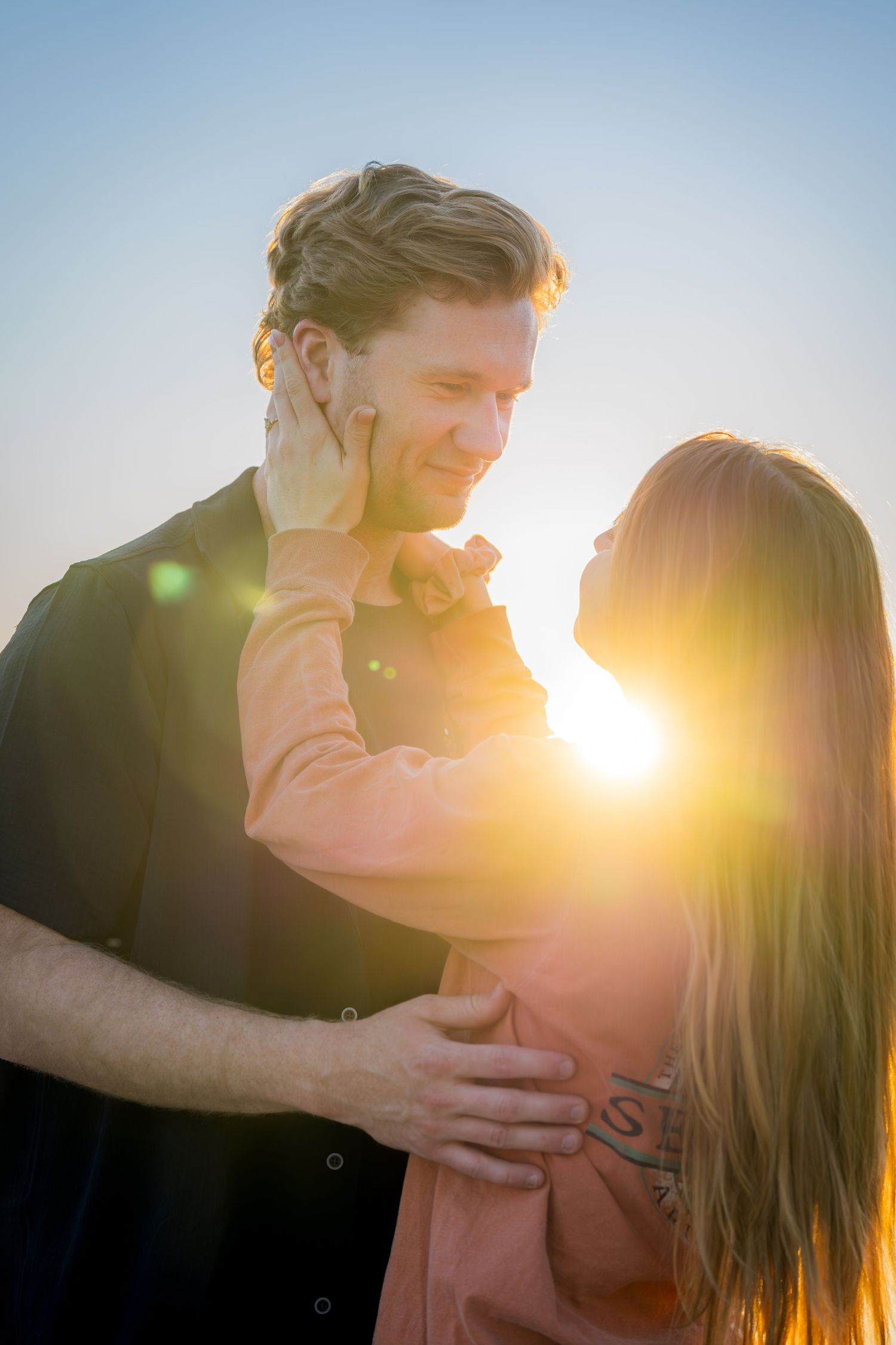 Romantic couple embracing at sunset with golden lens flare in a clear blue sky.