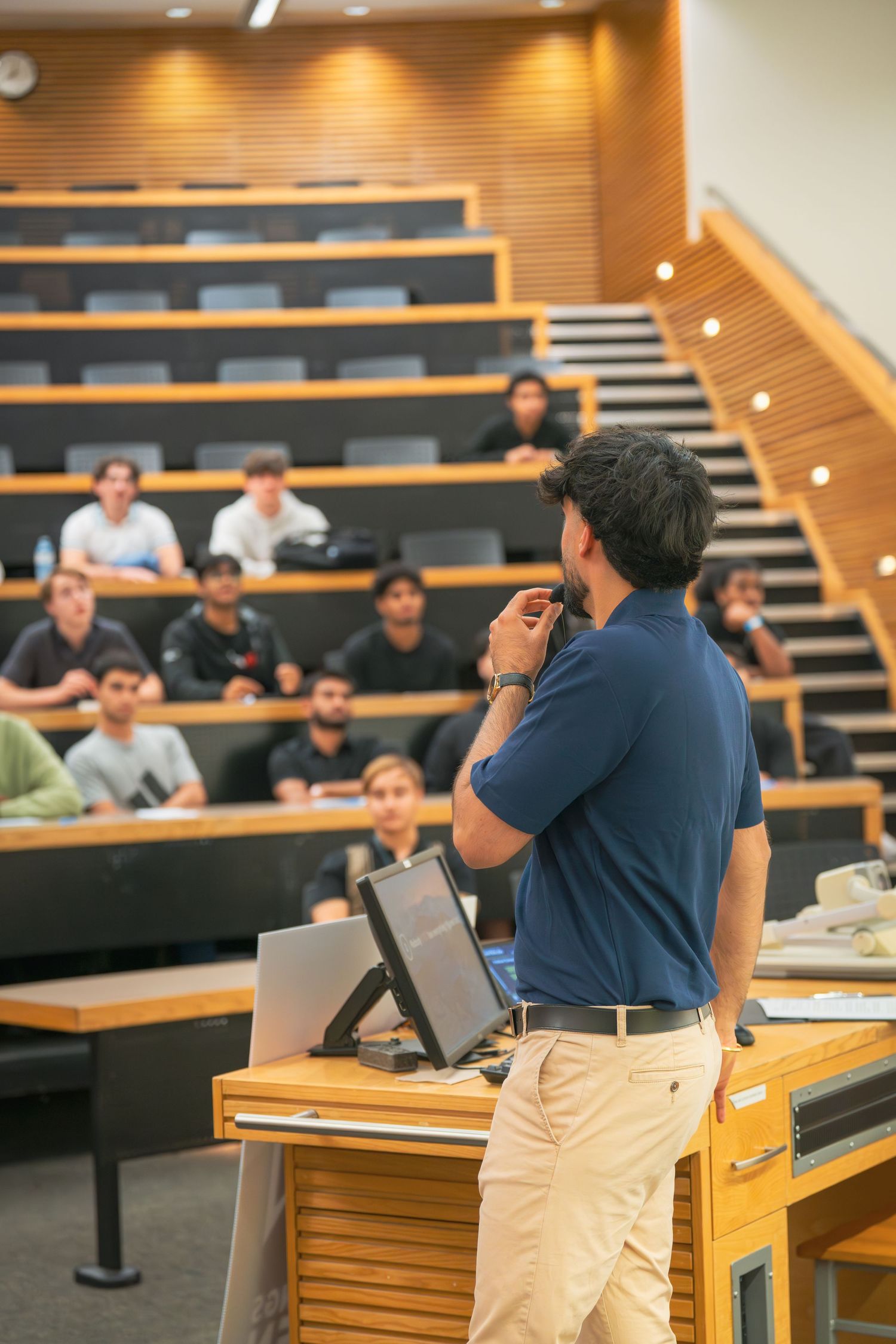 Presenter speaking at wooden podium in modern lecture hall with tiered seating.