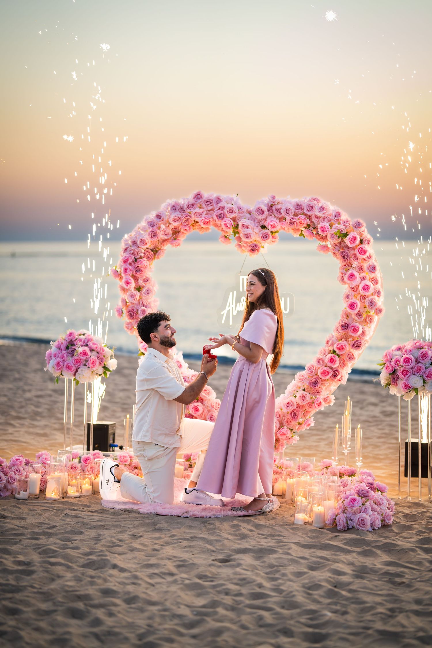 Marriage proposal scene on beach with illuminated heart arch and sparklers.
