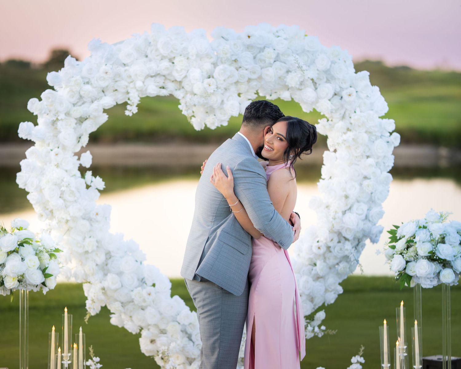 A romantic proposal setup with a white floral arch and glowing candles at sunset by a lake.