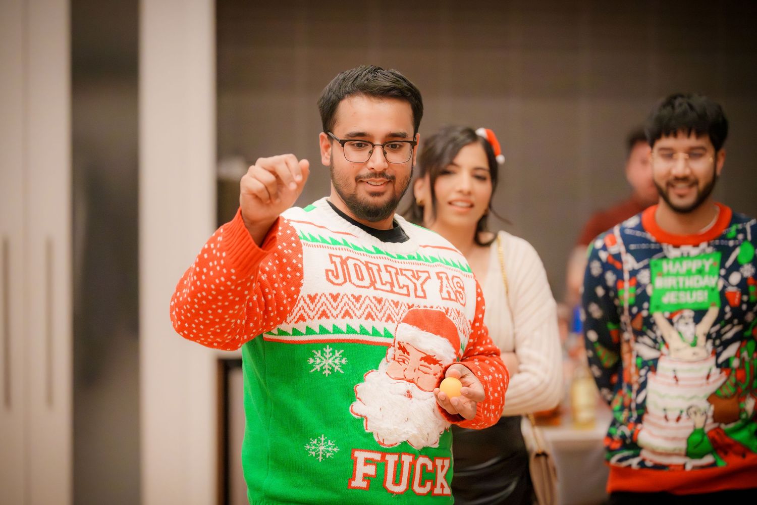 Person wearing a festive Christmas sweater with text at an indoor holiday party.