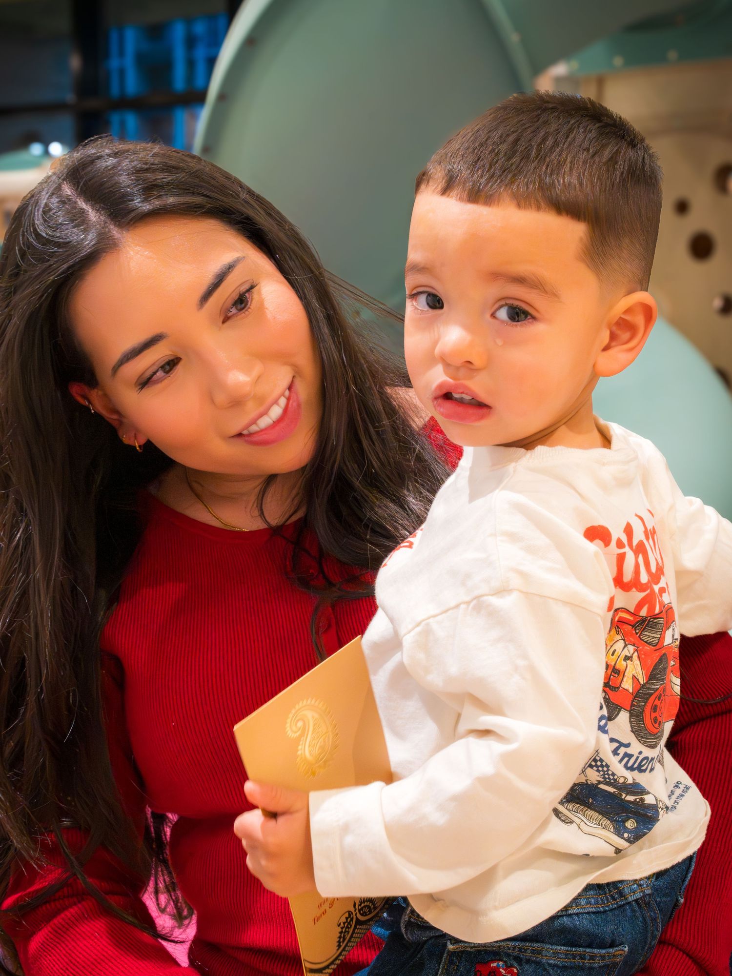 Young woman in red sweater holding a toddler boy at an indoor event.