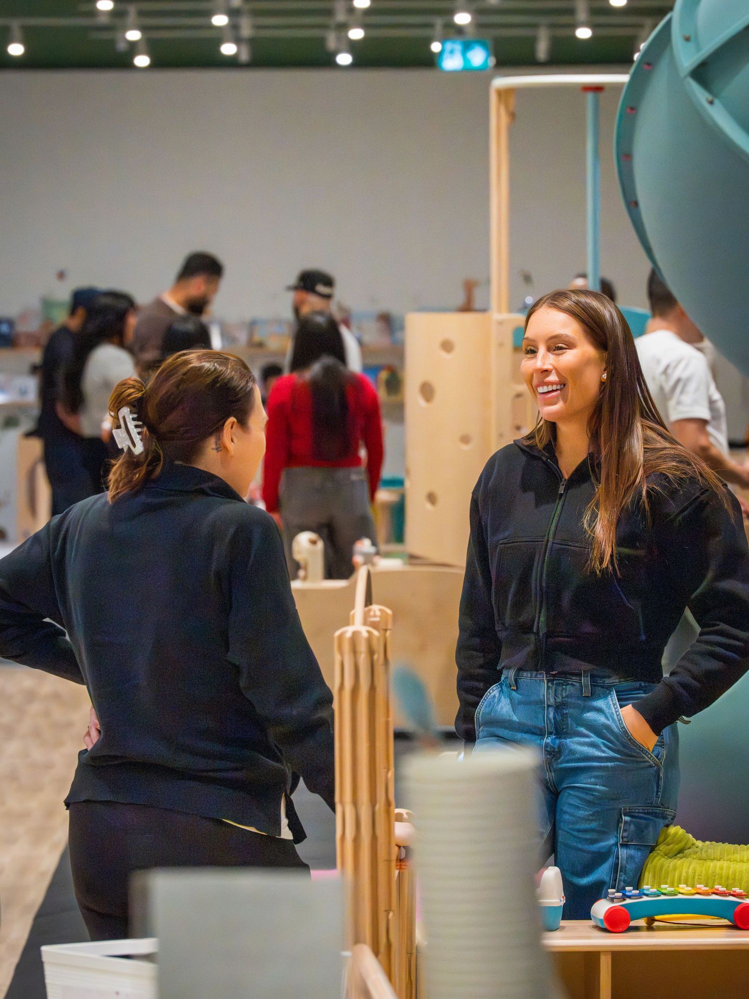 Two women in black outfits chat and smile in a bright open play space with families in background.