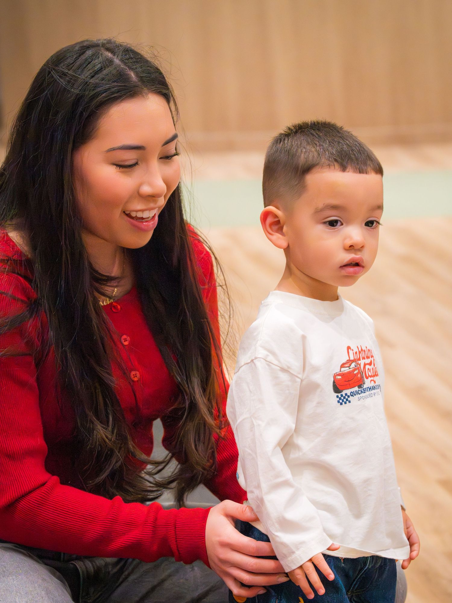 Young woman in red top smiling at toddler boy wearing Lightning McQueen shirt indoors.