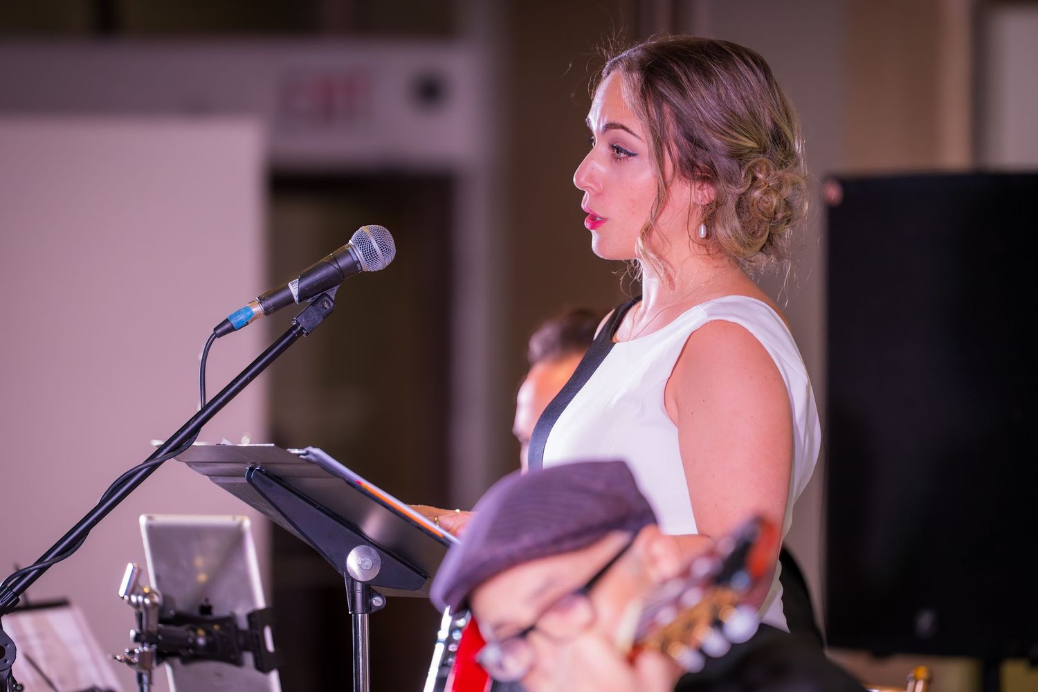 Blonde woman in white dress speaks at a microphone on stage during a live musical performance event.